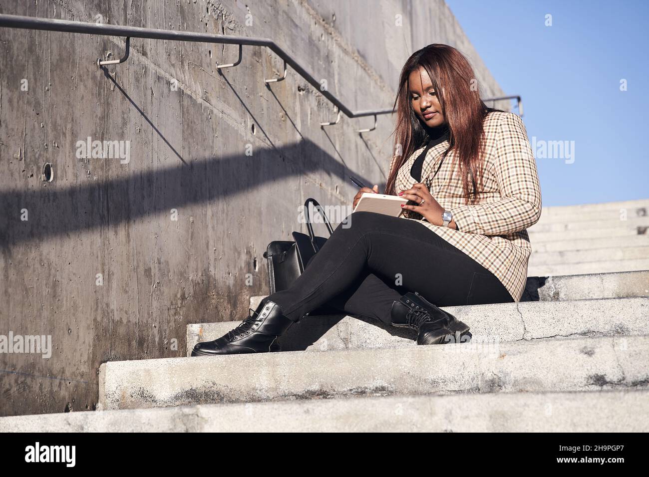 Young, African-American student sits outside on the university steps writing notes in a notebook. Concept of education Stock Photo