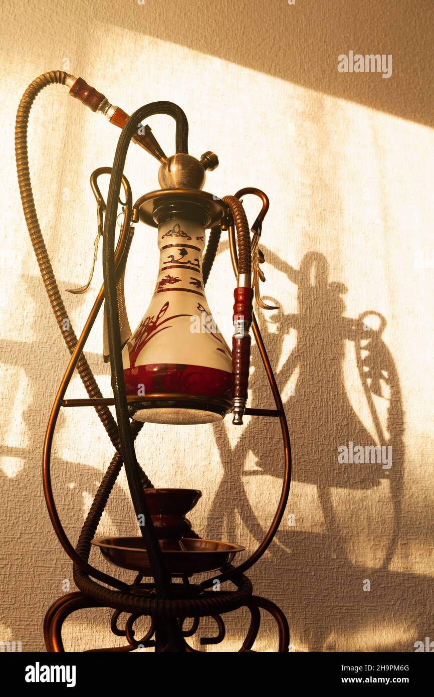 A hookah casting its shadow on a white stippled wall. Next to it is the shadow of another shisha. The light coming in is golden from the sun through a Stock Photo