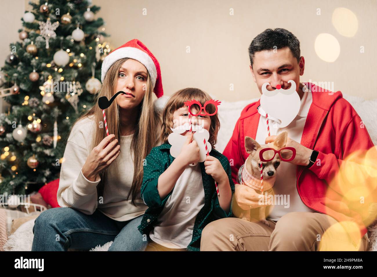 Portrait of happy family with kid son and puppy holding festive party props for photo booth. Mother in Santa hat, father, child boy and dog in sweater Stock Photo