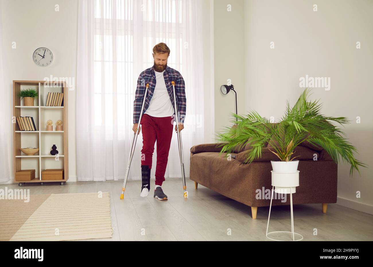 Young man with broken foot or leg wearing support brace and walking with crutches at home Stock Photo