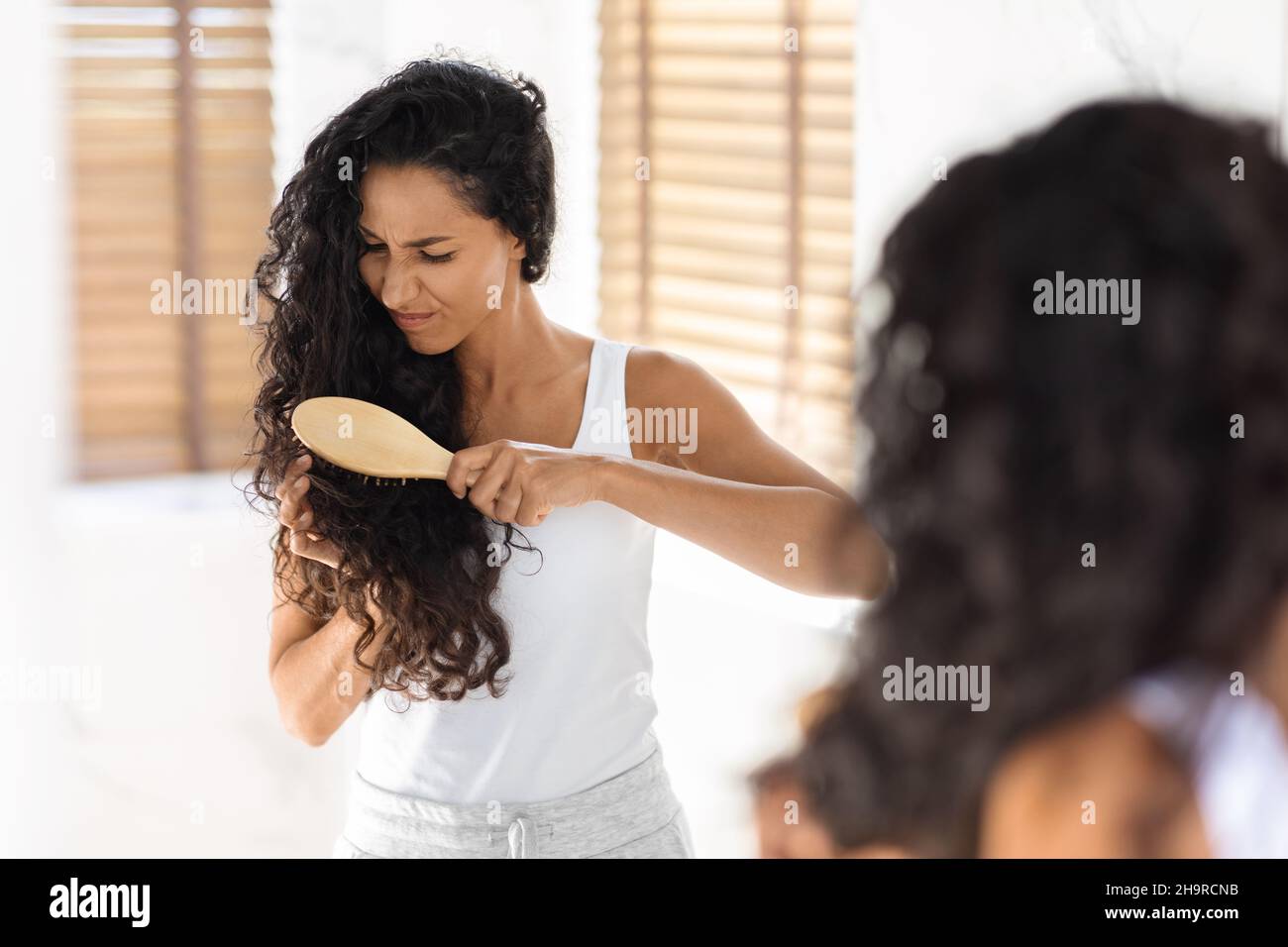 Young Brunette Woman Irritated By Tangled Hair, Using Bamboo Brush In Bathroom Stock Photo