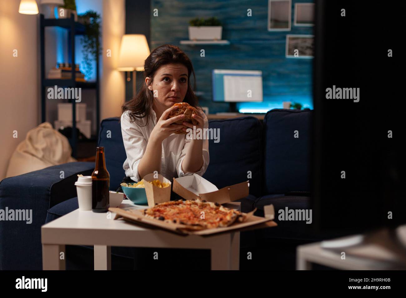 Woman sitting on sofa eating tasty delicious burger watching news on television in front of coffee table with large pizza. Caucasian female enjoying takeaway junk-food order. Stock Photo