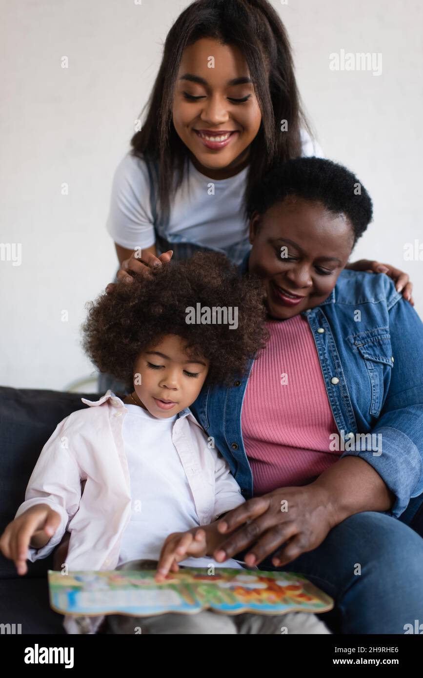 african american toddler girl looking at picture book near granny and happy mother Stock Photo