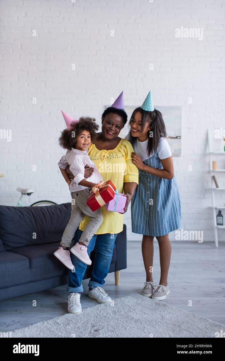 african american women and child in party caps holding presents while smiling at camera in living room Stock Photo