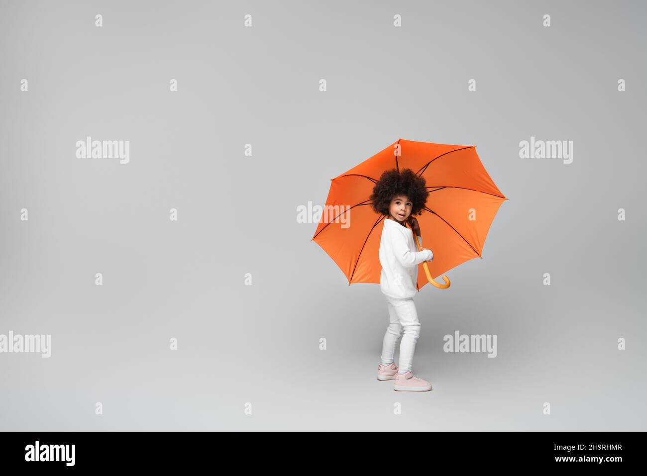 full length view of stylish african american girl under orange umbrella looking at camera on grey Stock Photo