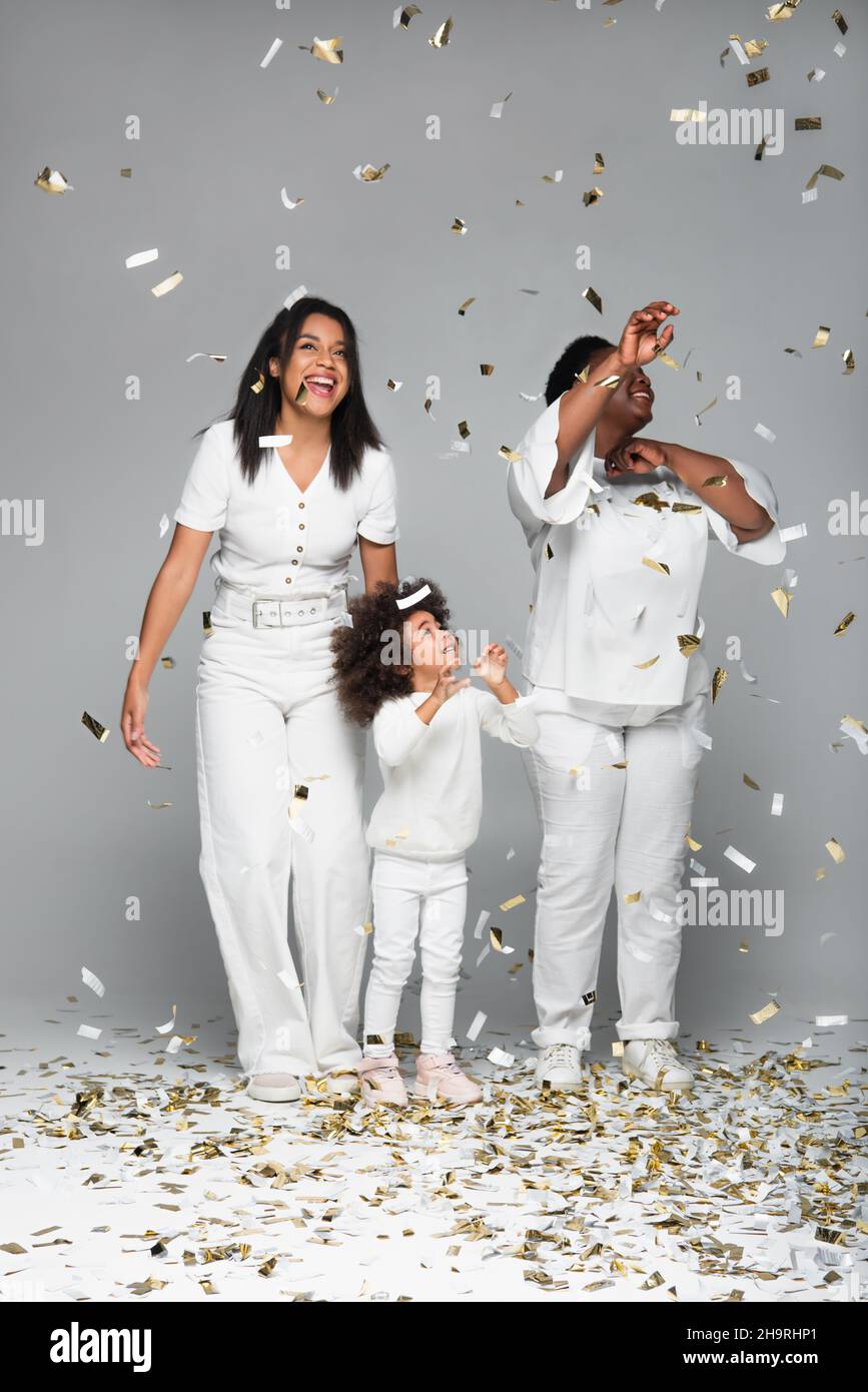 full length view of african american women and girl smiling under shiny confetti on grey Stock Photo