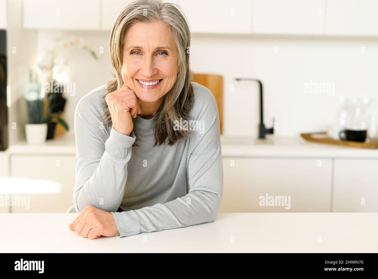 Elegant charming middle-aged woman with gray hair sitting at the countertop in bright modern kitchen and looks at the camera, smiling, put her chin on hands. Wellbeing concept Stock Photo
