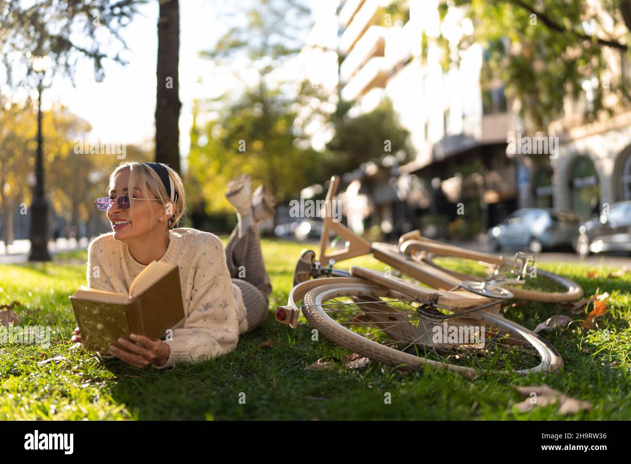 Positive female in sunglasses lying on grassy ground near timber bicycle and reading book in park Stock Photo