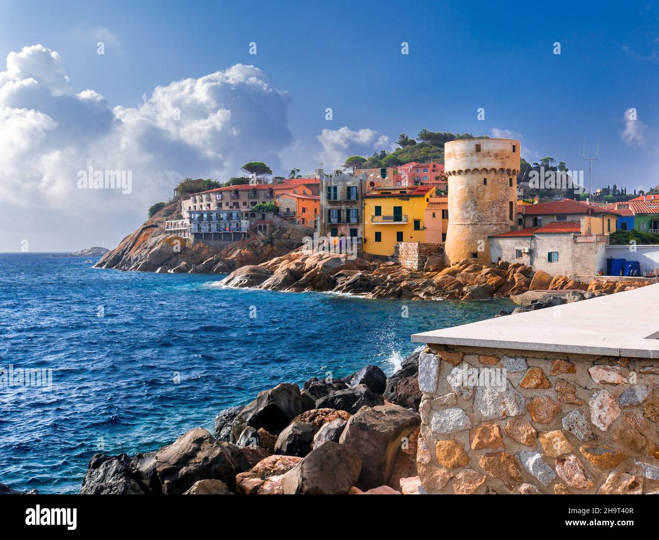 The perfect tiny seaside village of 'Giglio Porto' with multi colored houses, an ancient defensive tower and a rocky coastline against a deep blue Med Stock Photo