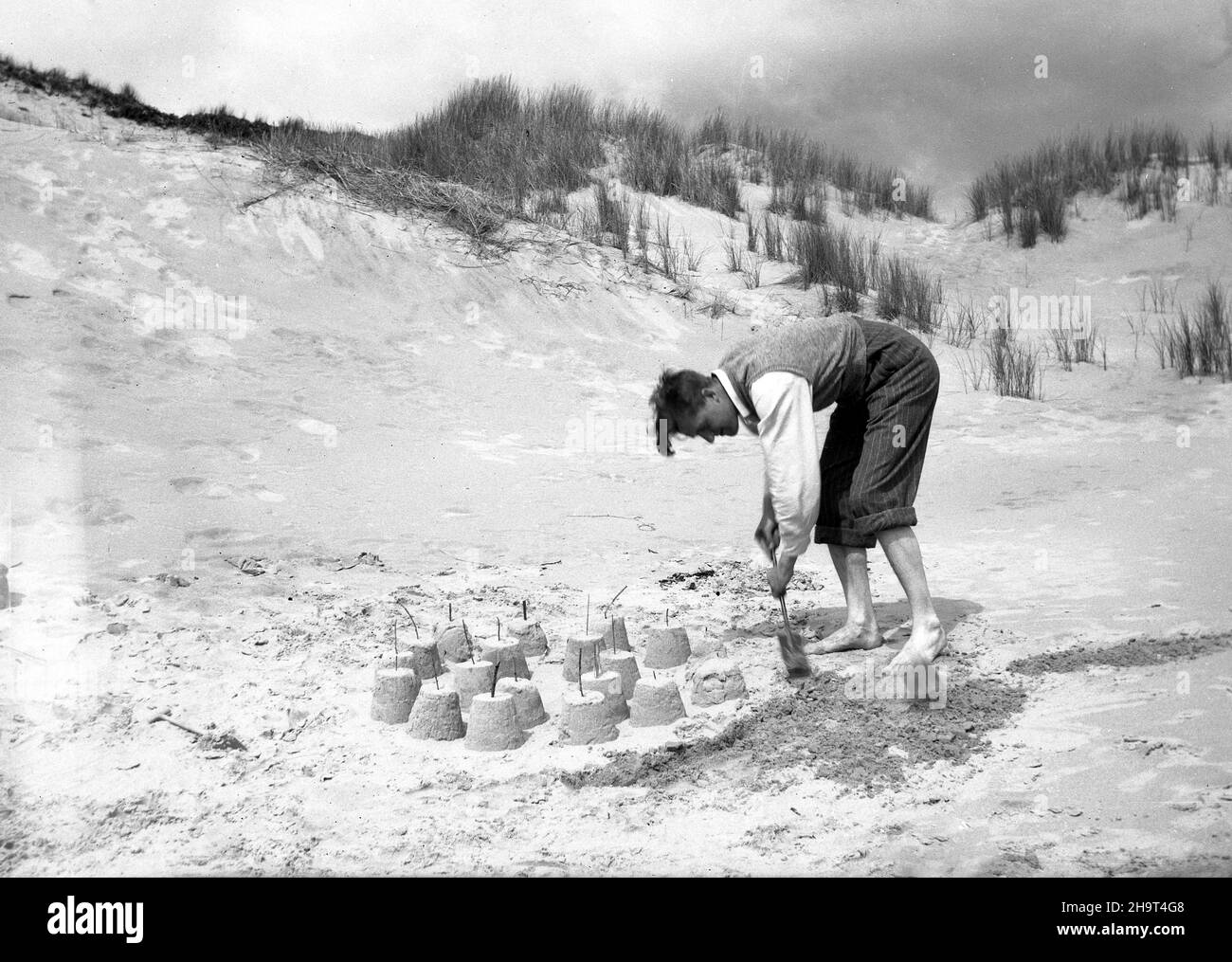 1950s, historical, outside on a sandy beach, by some sand dunes, a man with his pin stripe suit trousers rolled up to his knees and a sleeveless sweater over his shirt, ending down with a small child's spade, building sandcastles, all with little sticks placed on the top of them, England, UK. Stock Photo