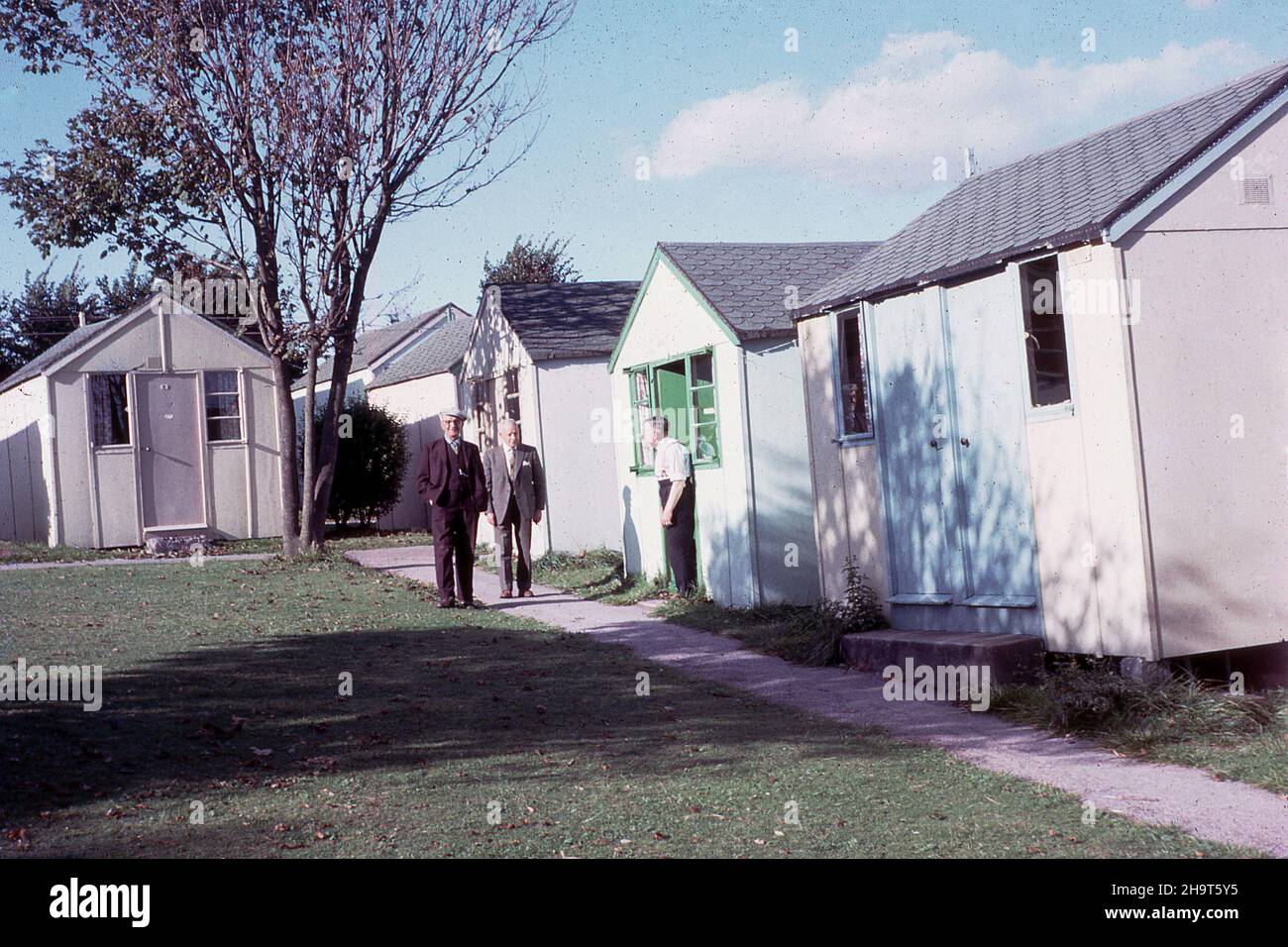 1964, historical, two gentlemen outside on a path and another gentleman standing a the entrance to his chalet at a Pontin's holiday camp, England, UK. Founded by Fred Pontin in 1946, the camps offered affordable holidays for the working man and his family, with basic accomodation in small single-storey pre-fab huts or chalets and on-site dining, sports activities and entertainment. The British holiday camp industry boomed from the end of WW2 to the 60s, but in the 1970s the rapid growth of the overseas package holiday saw it decline and in the 1980s many sites closed. Stock Photo