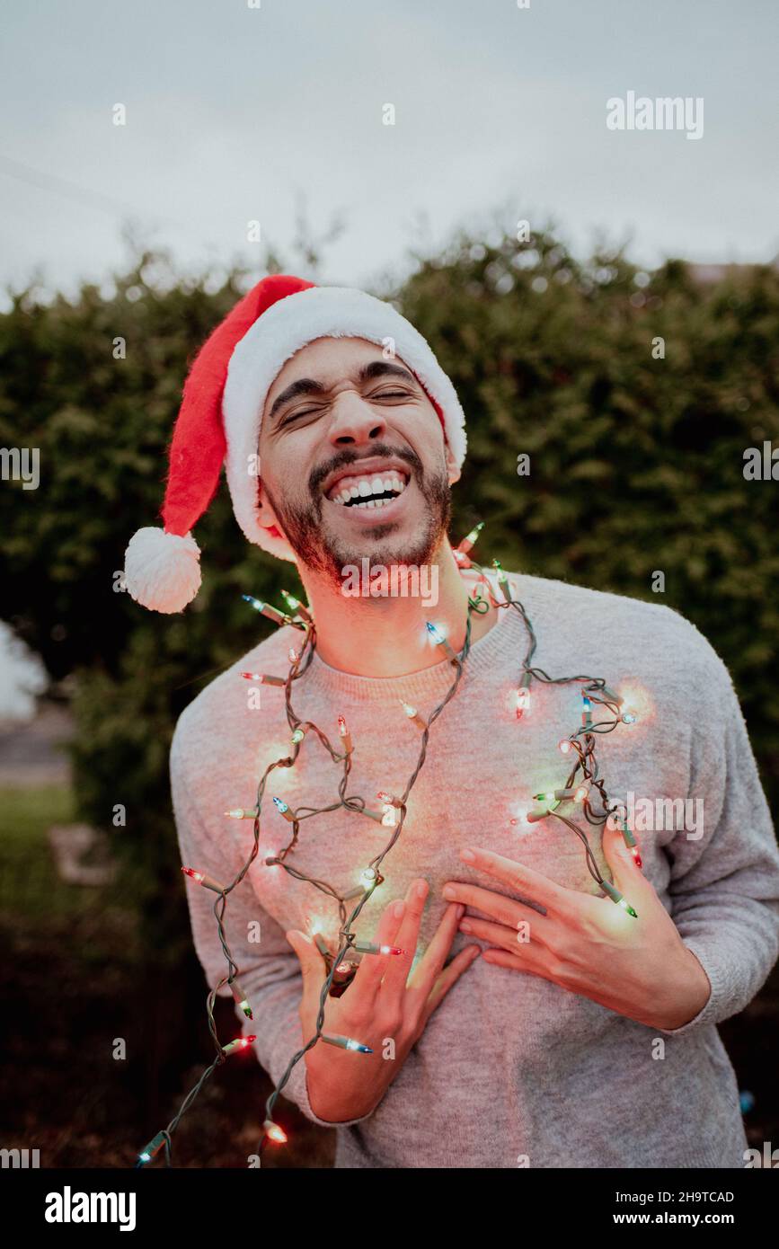 Mixed-race man laughs in santa hat and sweater wearing holiday lights around his neck in the winter outside in front of evergreen tree Stock Photo