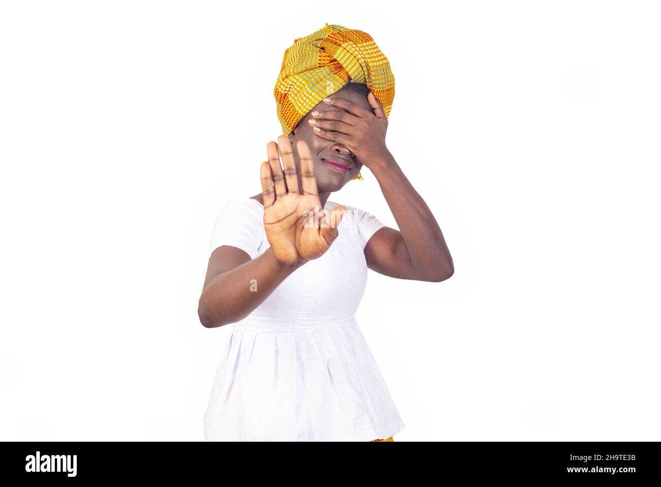 beautiful young african woman in traditional dress standing on white background waving hitch and covering her face with hand. Stock Photo