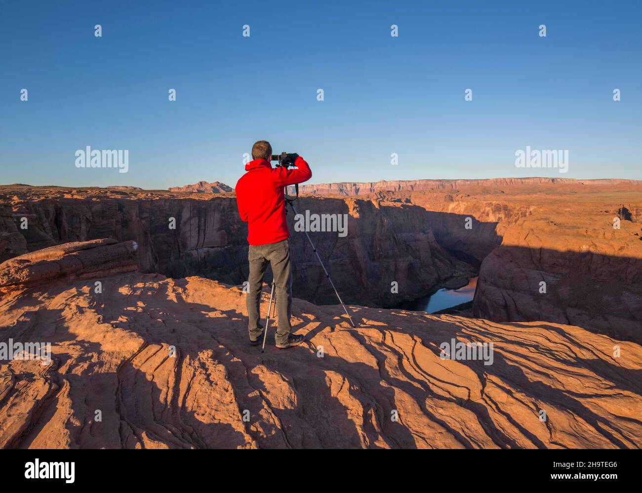 Glen Canyon National Recreation Area, Page, Arizona, USA. Photographer at cliff edge shooting view over the Colorado River at Horseshoe Bend, sunrise. Stock Photo
