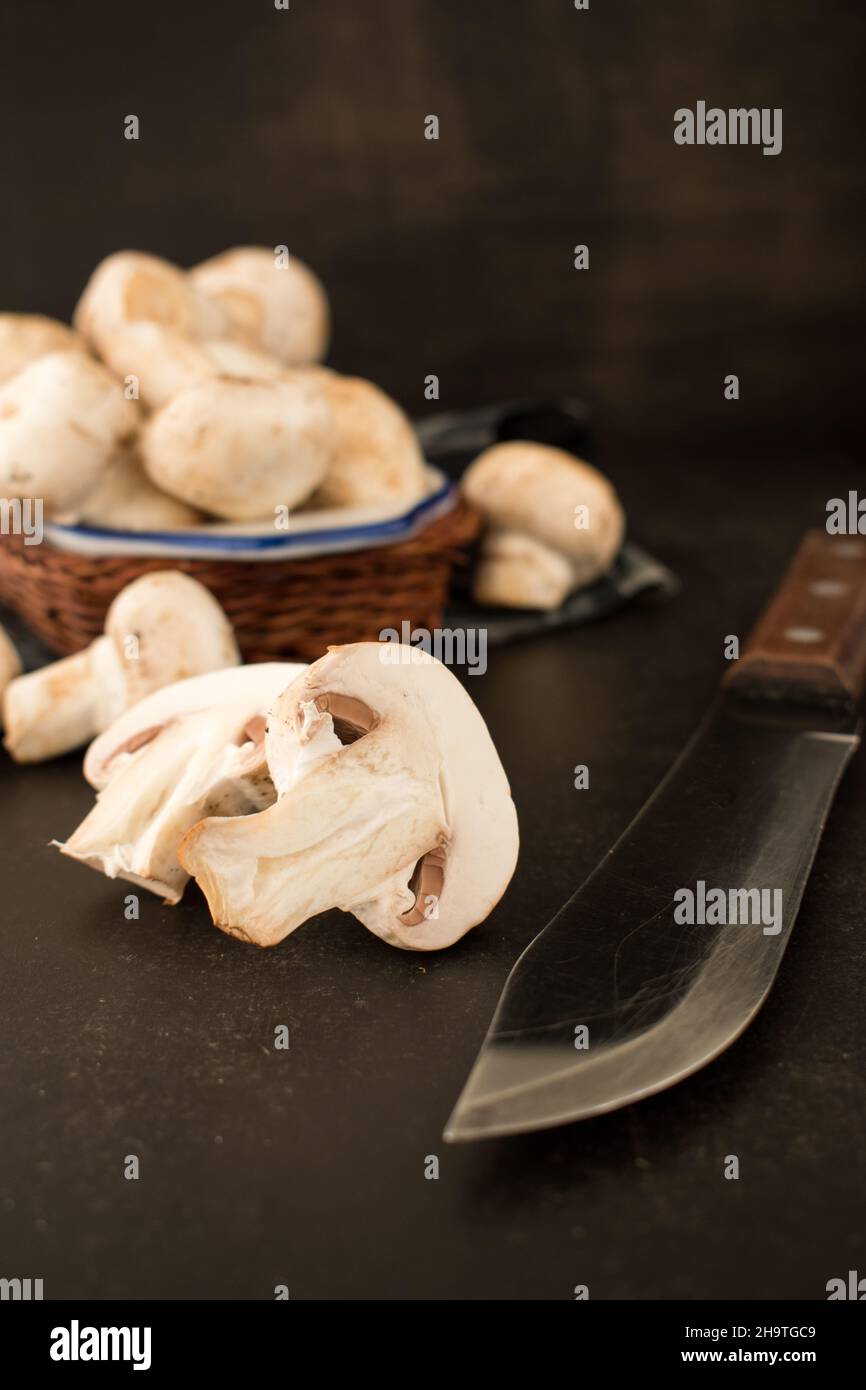 Cut champignon and some mushrooms in a basket with a knife on a black wooden table Stock Photo