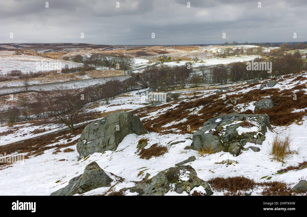 North York Moors with view of rocky boulders flanked by heather, grasses, and trees under cloudy sky and landscape following snowfall. Goathland, UK. Stock Photo