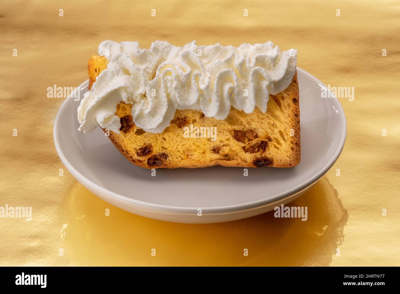 Slice of panettone cake with whipped cream in white plate on a golden background, typical Italian Christmas dessert Stock Photo