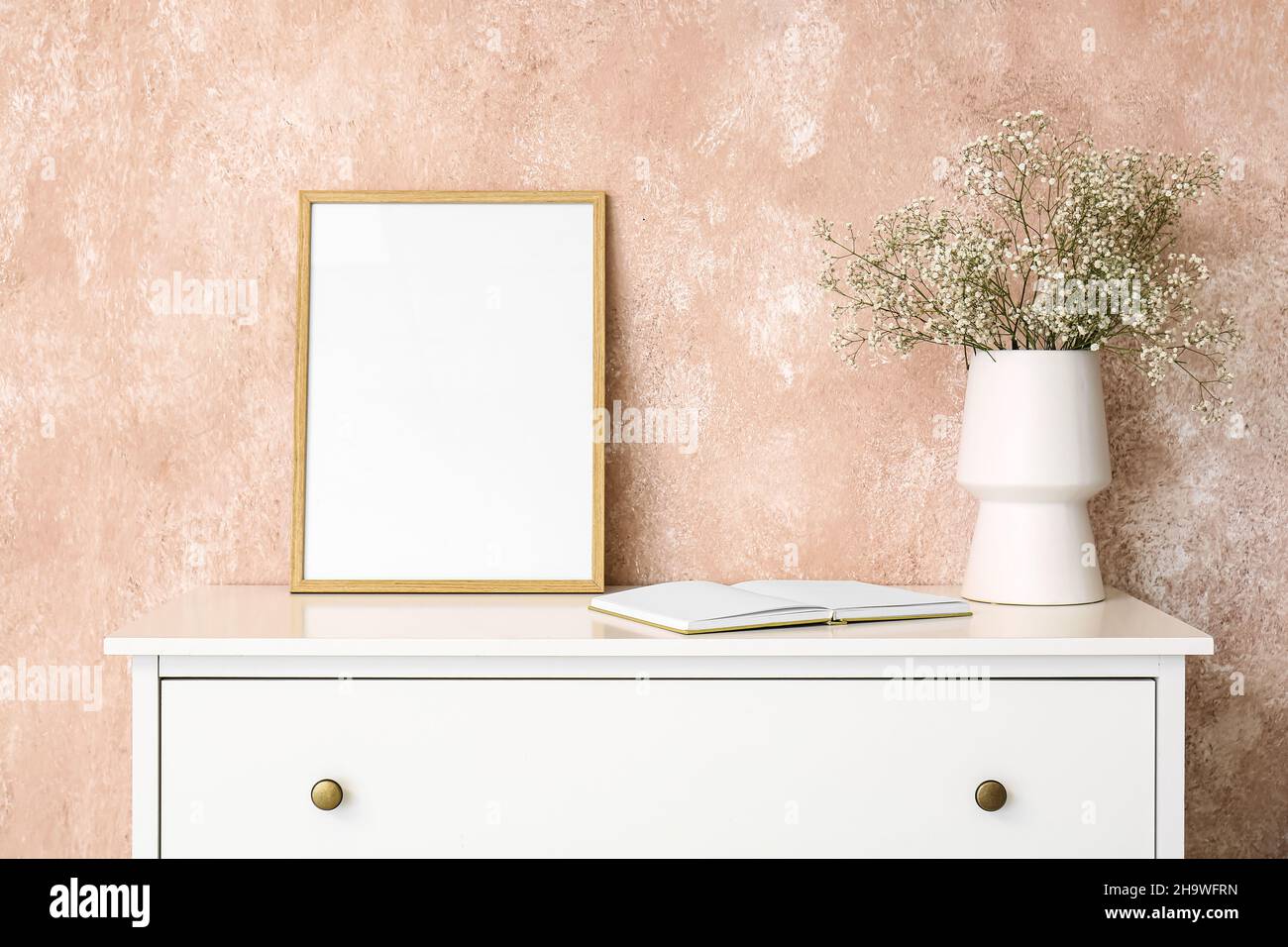 Vase with gypsophila flowers, book and frame on shelf near pink wall ...
