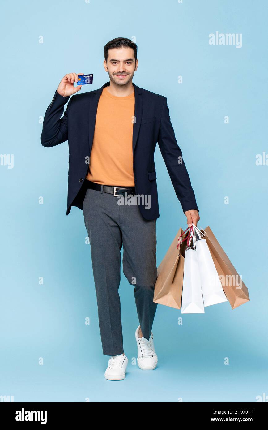 Full lenght portrait of young smiling Caucasian man holding shopping bags and showing credit card in isolated light blue studio background Stock Photo