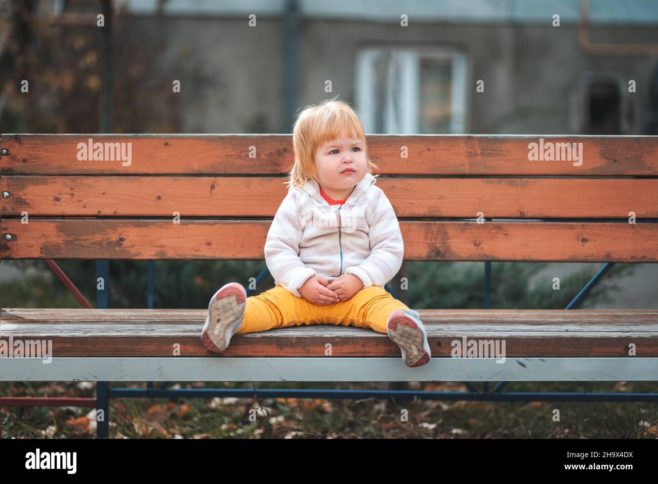 Childrens shelter. A sad little girl is sitting on a bench on the playground alone. The concept of lost childrens. Stock Photo