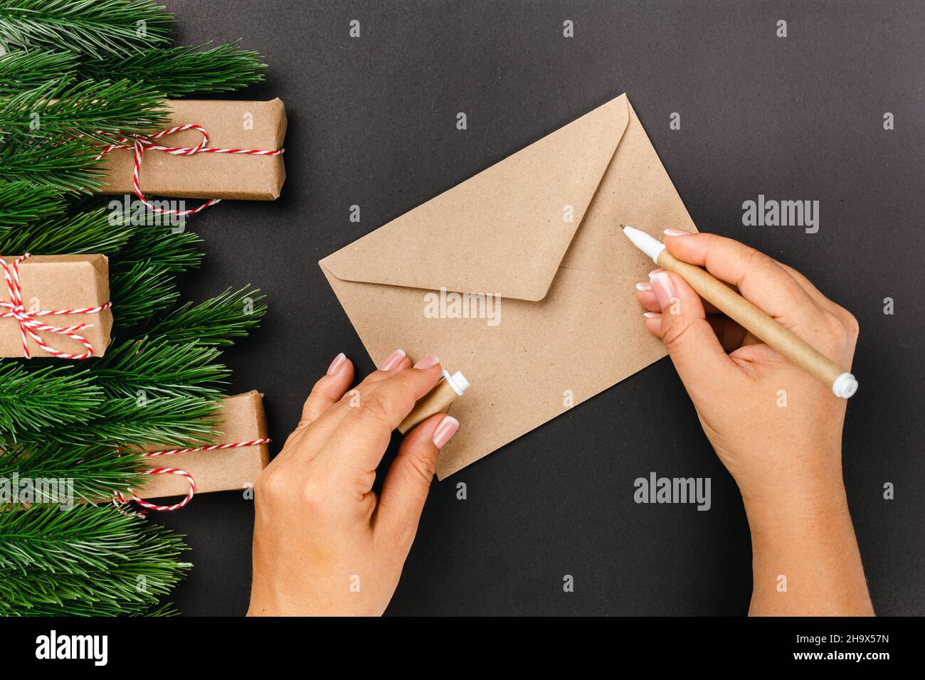 flat lay on woman hands writing letter in a Santa Claus craft envelope on black background with frame made of Christmas tree branches Stock Photo