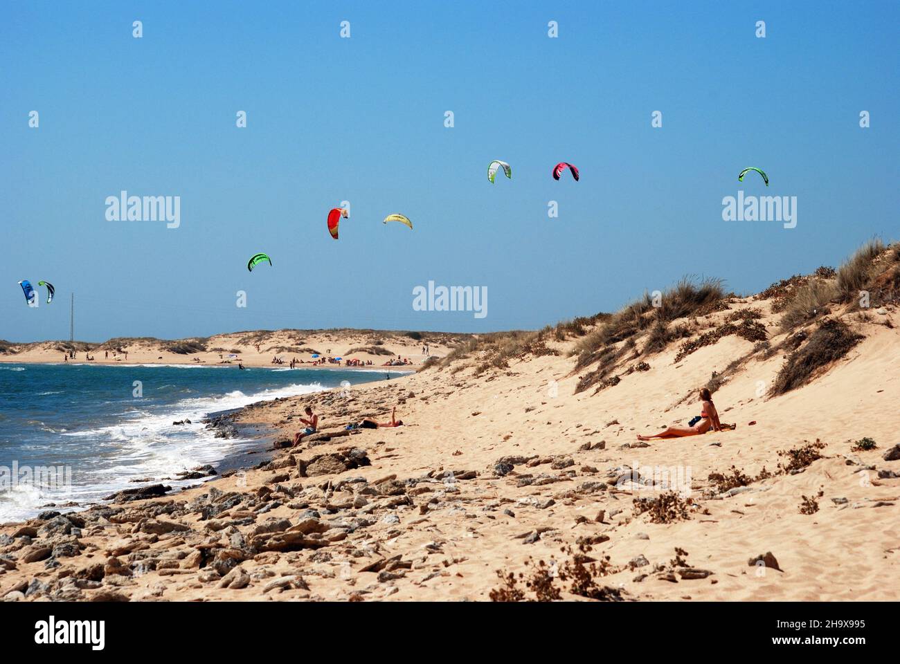 Tourists relaxing on the beach with kitesurfers to the rear, Cabo Trafalgar, Cadiz Province, Andalusia, Spain. Stock Photo