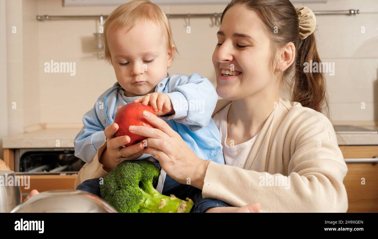 Young mother showing kitchen cookware, utensils and ingredients to her baby son on kitchen