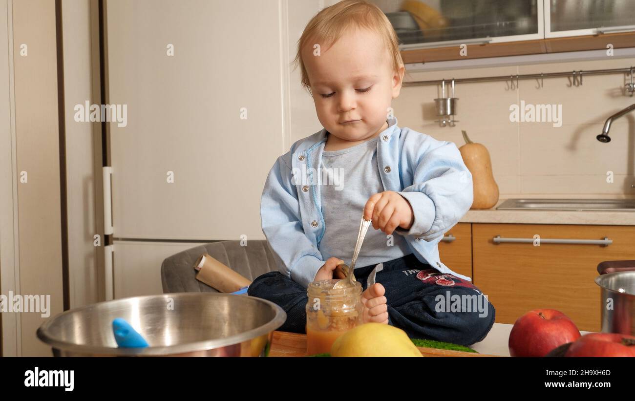 Little 1 year old baby boy sitting on kitchen table and eating fruit