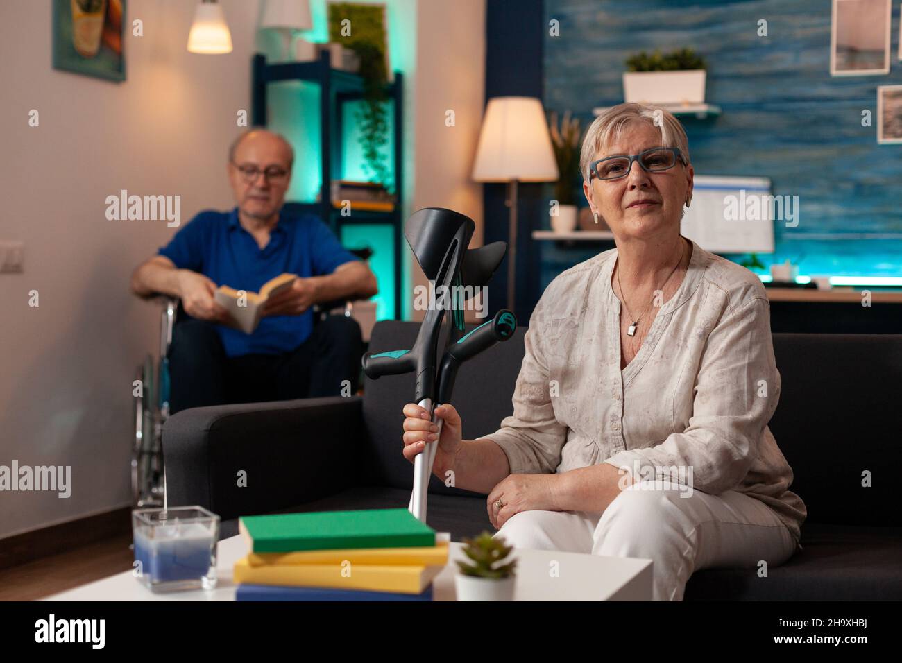 Portrait of aged woman sitting on couch and looking at camera while senior man in wheelchair reading book in background. Old person holding crutches on sofa with disabled husband in living room Stock Photo