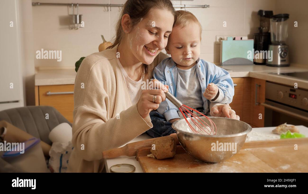 Little baby boy looking a this smiling mother kneading dough in bowl on ...