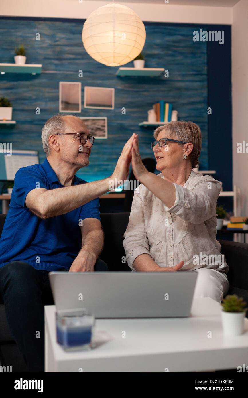 Retired man and woman highfive while using laptop on table at home. Senior married couple enjoying time together with technology while sitting on living room sofa. Old cheerful people Stock Photo