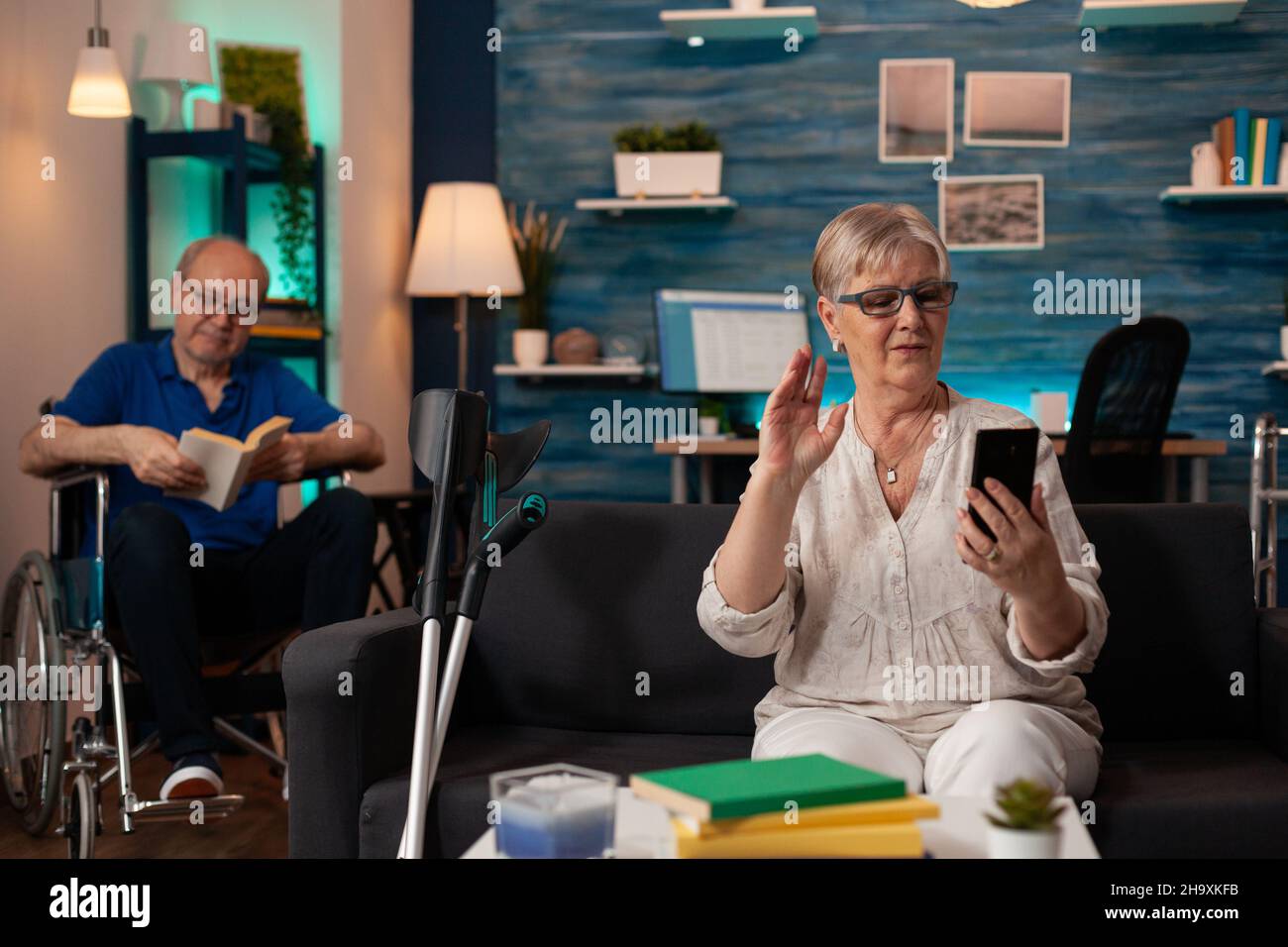 Caucasian old woman talking on video call with smartphone on living room sofa. Person using online remote conference while disabled senior man sitting in wheelchair with book in background Stock Photo