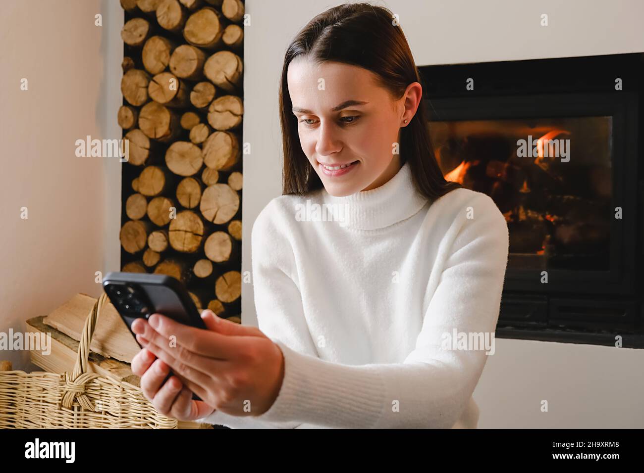 Attractive young millennial lady sitting on warm floor relaxing inside of living room of country cottage house near fireplace spend time using phone Stock Photo
