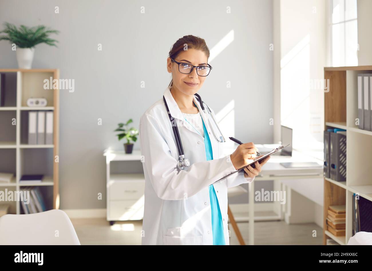 Portrait of young female doctor standing with clipboard in hands at modern medical center office. Stock Photo