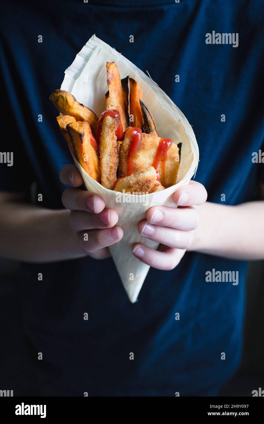 A child holding a mini escalope, sweet potato fries and ketchup in a bamboo cone Stock Photo