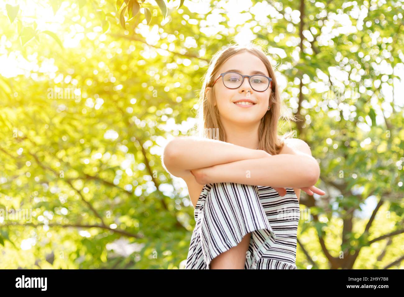 Portrait of a smiling girl wearing glasses with her arms crossed over her knee on a sunny wooded background Stock Photo