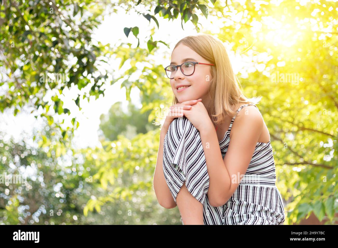Portrait of a smiling girl with glasses looking to the side with her arms crossed over her knee on a sunny wooded background Stock Photo