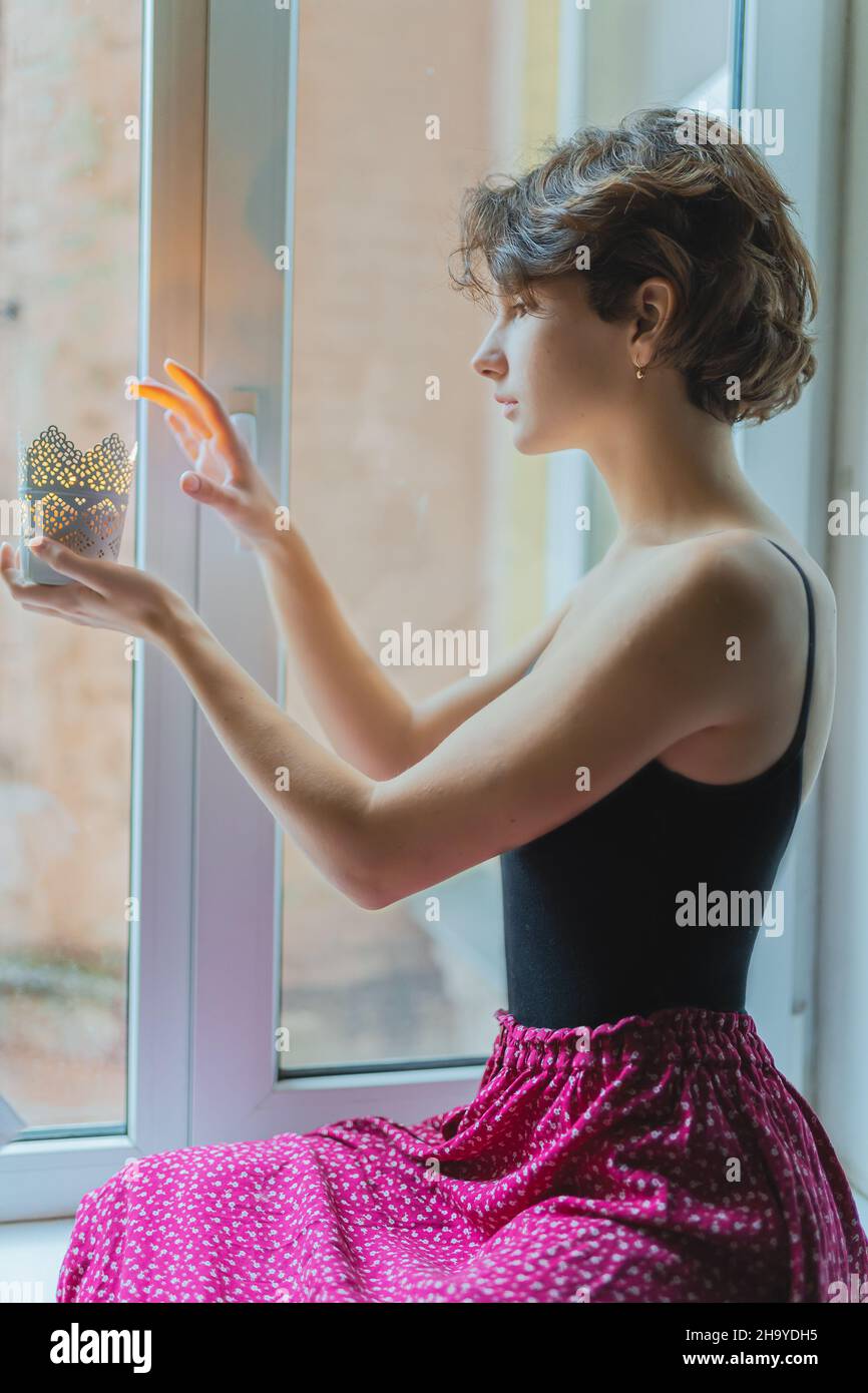 Silhouette of beautiful young lady with brown curly hair in authentic clothes sitting on windowsill. The girl's hands are holding candlestick Stock Photo