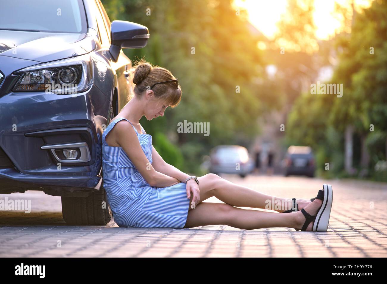 Young woman driver sitting beside her broken car waiting for help. Vehicle problems concept. Stock Photo