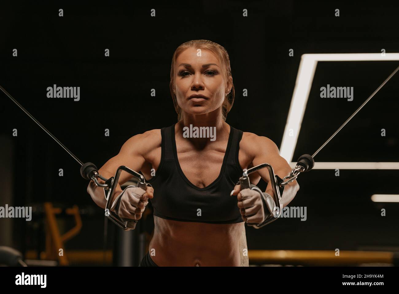 A close-up photo of a fit woman with blonde hair who is doing a chest  workout on the cable machine in a gym. A girl is training her pectoral  muscles Stock Photo 