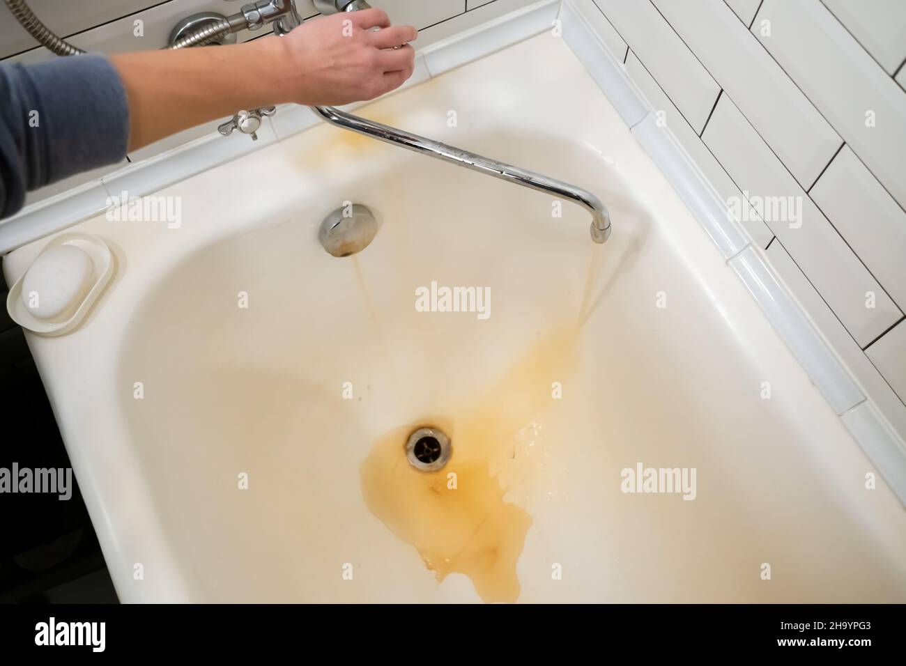 Female hand opens a tap, from which rusted water flows into a white bath. Problem is old pipes that need to be replaced. Stock Photo
