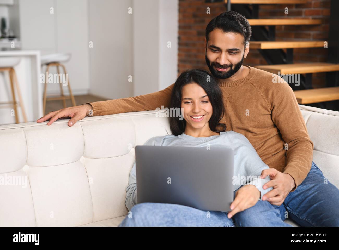 Young Pakistani couple using laptop while sitting on the sofa at home. Spouses watching movie on laptop. Happy man and woman embracing while relaxing on couch Stock Photo