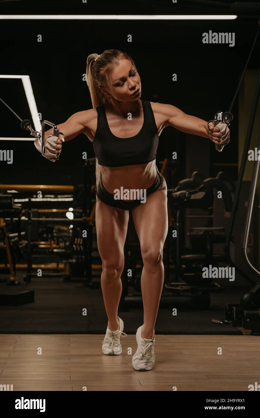 A close-up photo of a fit woman with blonde hair who is doing a chest  workout on the cable machine in a gym. A girl is training her pectoral  muscles Stock Photo 