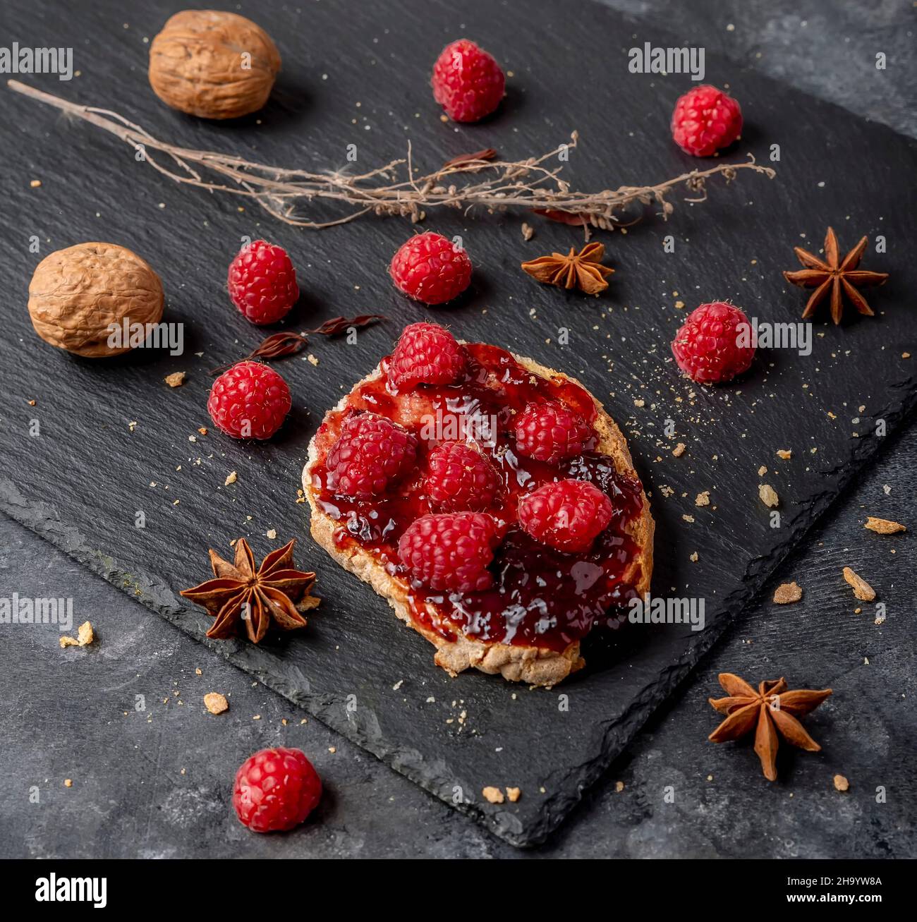 A delicious crouton with jam and whole raspberries on top and some walnuts near it, on a black slate background Stock Photo