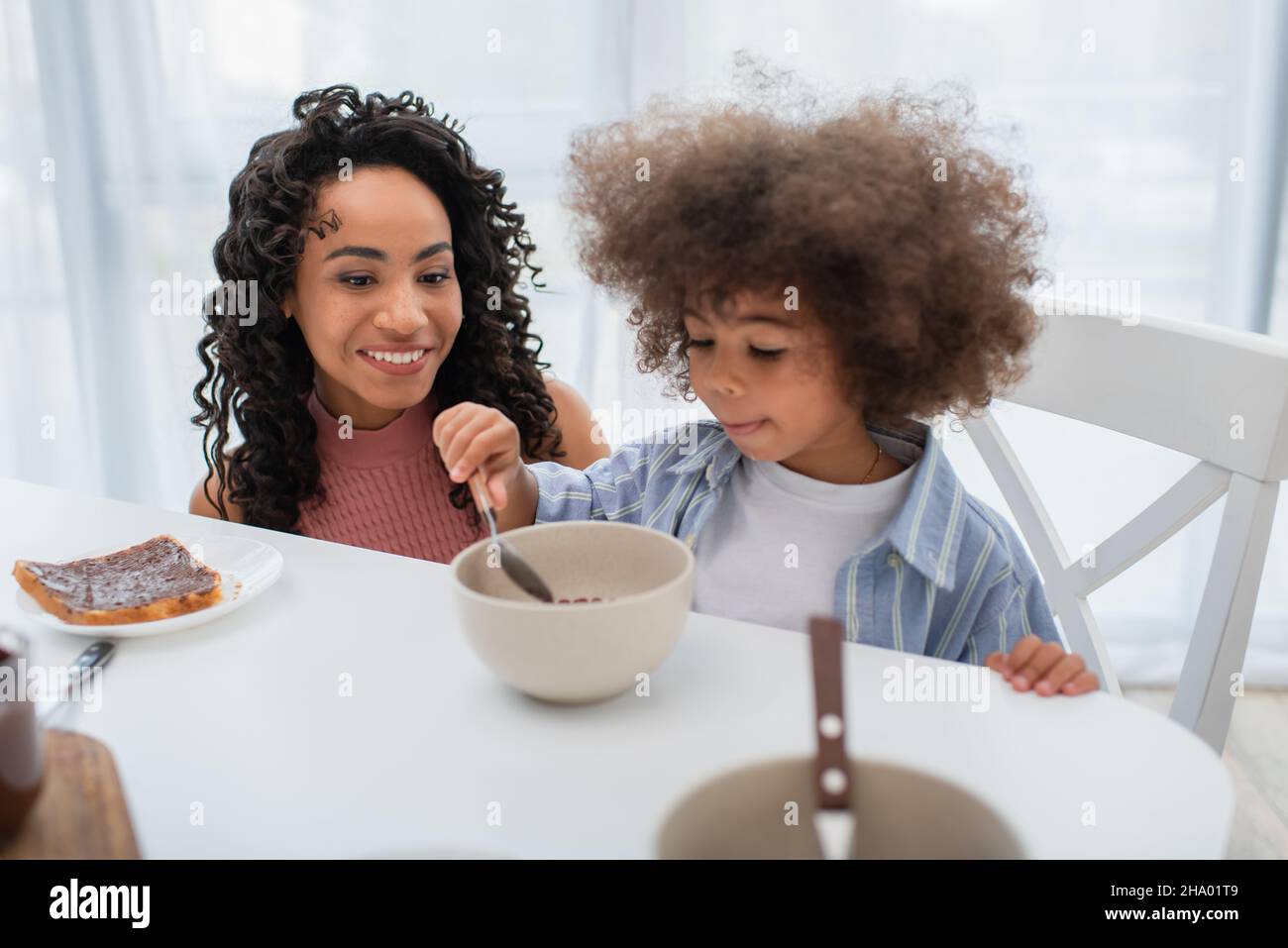 Happy african american woman looking at daughter with cereal in kitchen Stock Photo