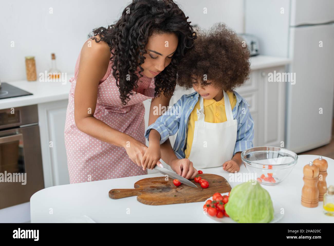 Young african american woman cutting cherry tomatoes near daughter and cabbage in kitchen Stock Photo