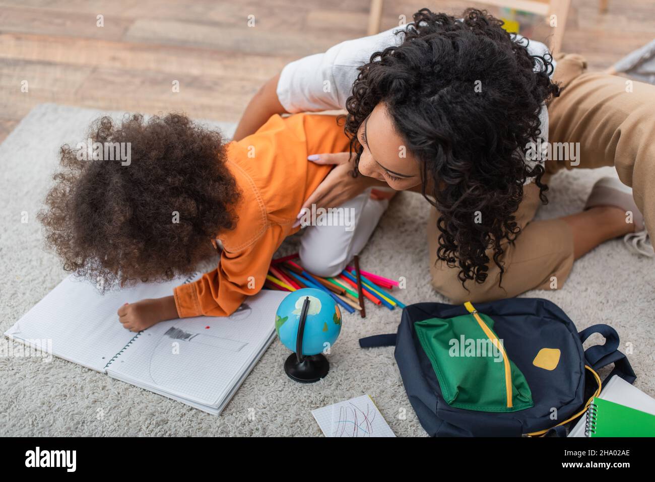 African american mother hugging child drawing on notebook near globe and backpack Stock Photo