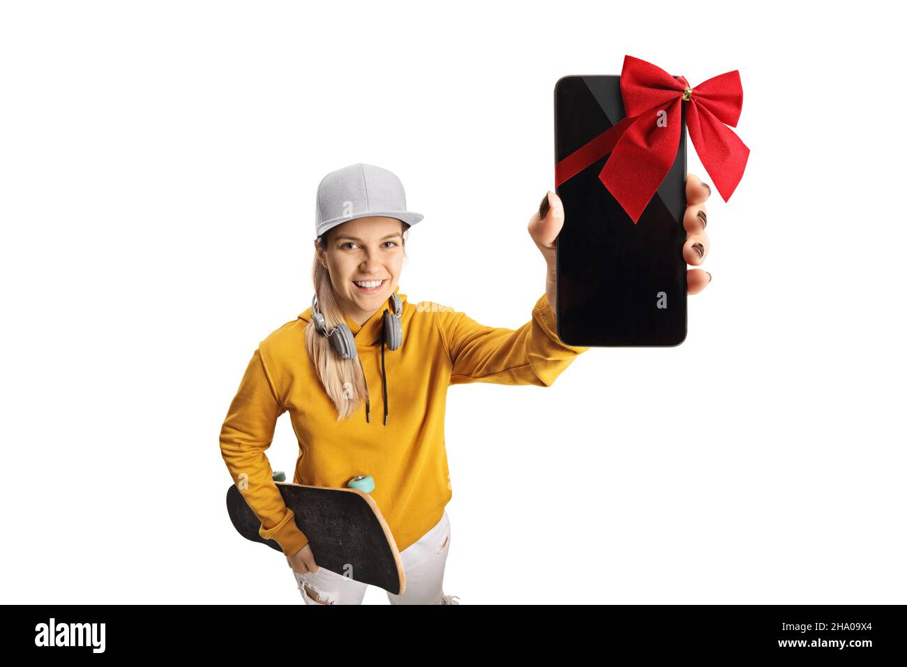 Female teen with headphones and a skateboard holding a smartphone with red bow in front of camera isolated on white background Stock Photo