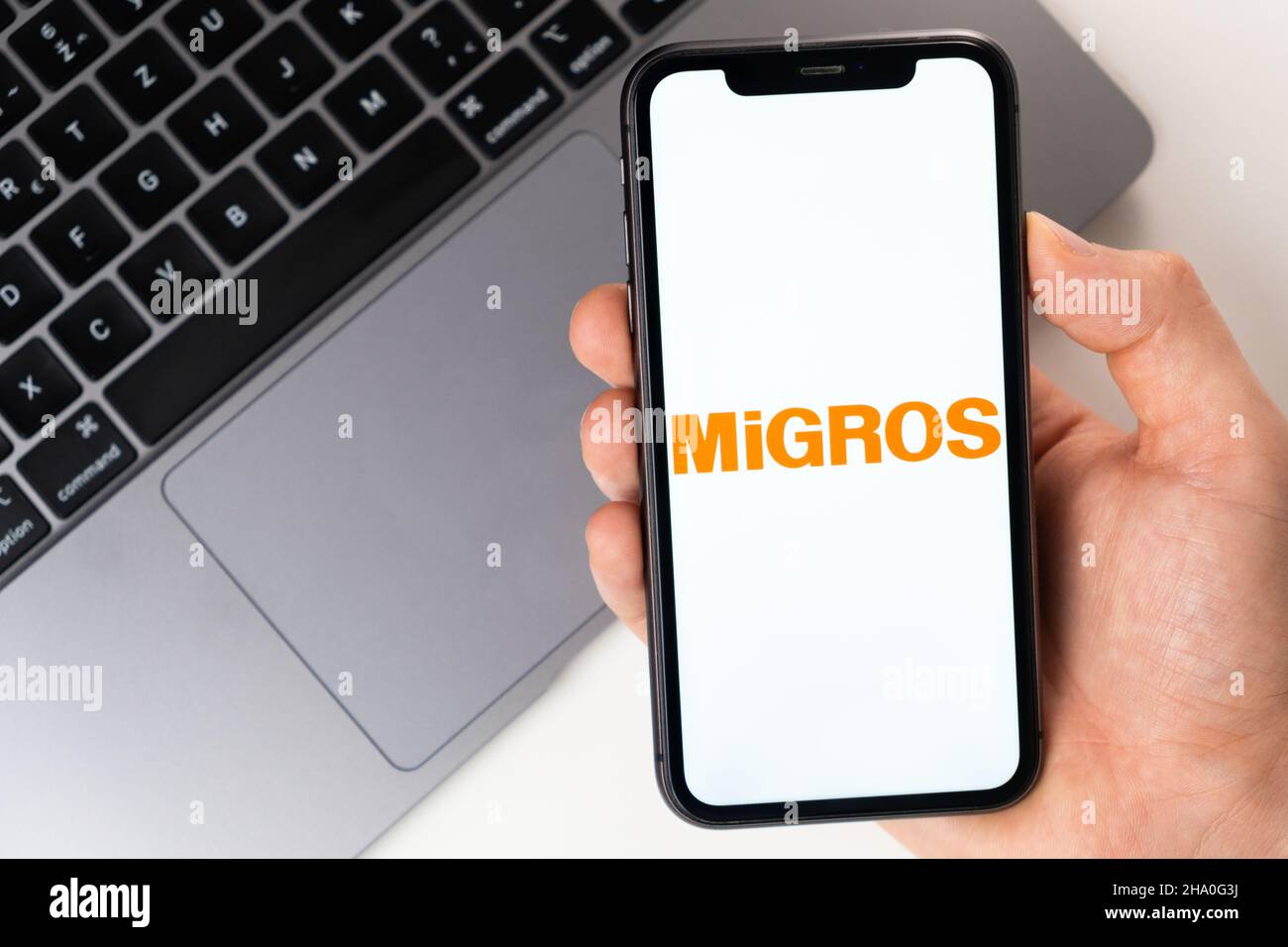 MiGROS mobile application on the smartphone screen. A mobile phone in a man hand near an open laptop. White background. November 2021, San Francisco, USA Stock Photo