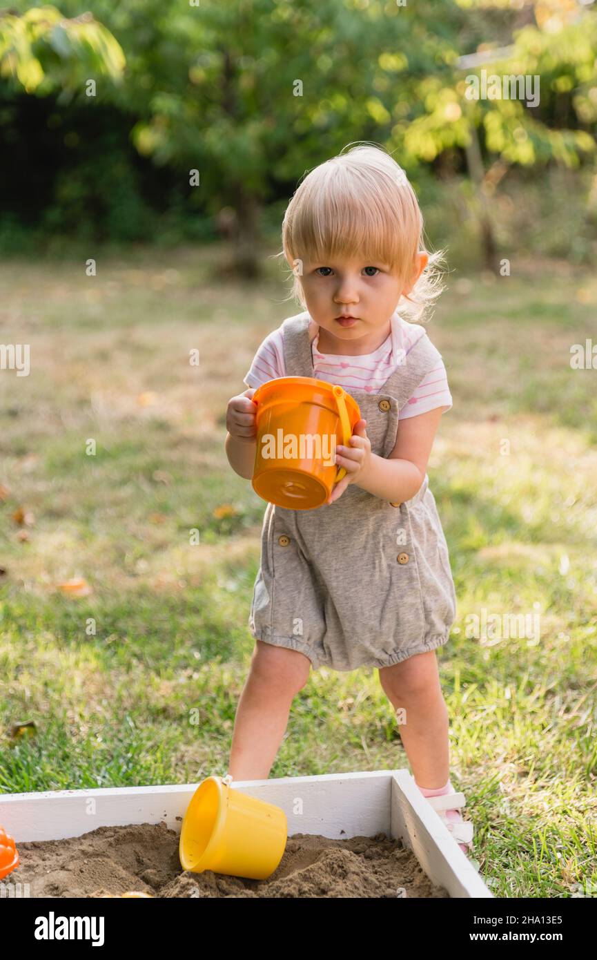 A little serious girl in a gray jumpsuit with shorts with an orange toy bucket with sand in her hands is playing in the garden. There are other colorf Stock Photo